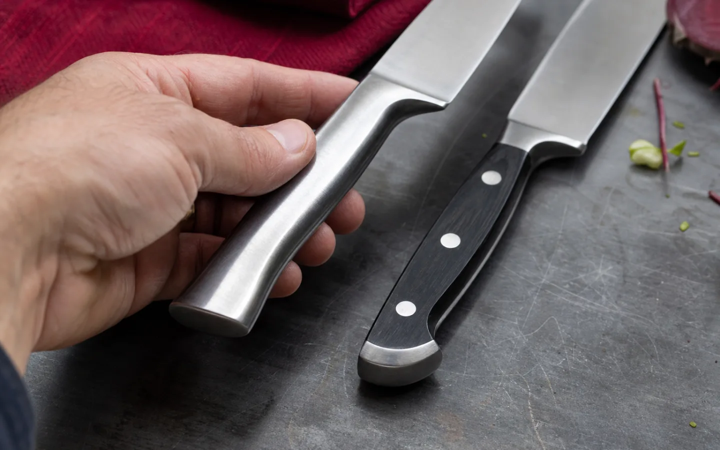 En hand som håller en kockkniv med stålhandtag bredvid två knivar med mörka, nitade handtag